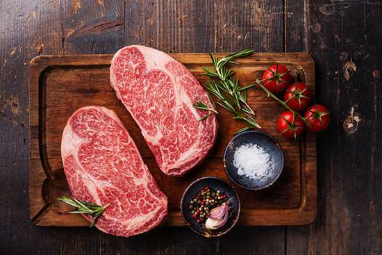 Two well-marbled ribeye steaks on a cutting board with tomatoes, herbs, and spices.