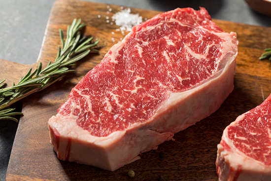 A raw New York strip steak with rosemary on a wooden cutting board.