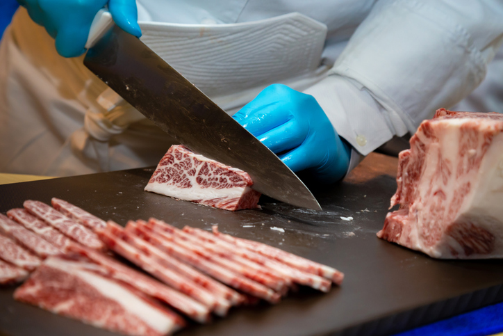 A meat cutter wearing blue gloves slicing some raw beef marbled with fat into thin pieces.