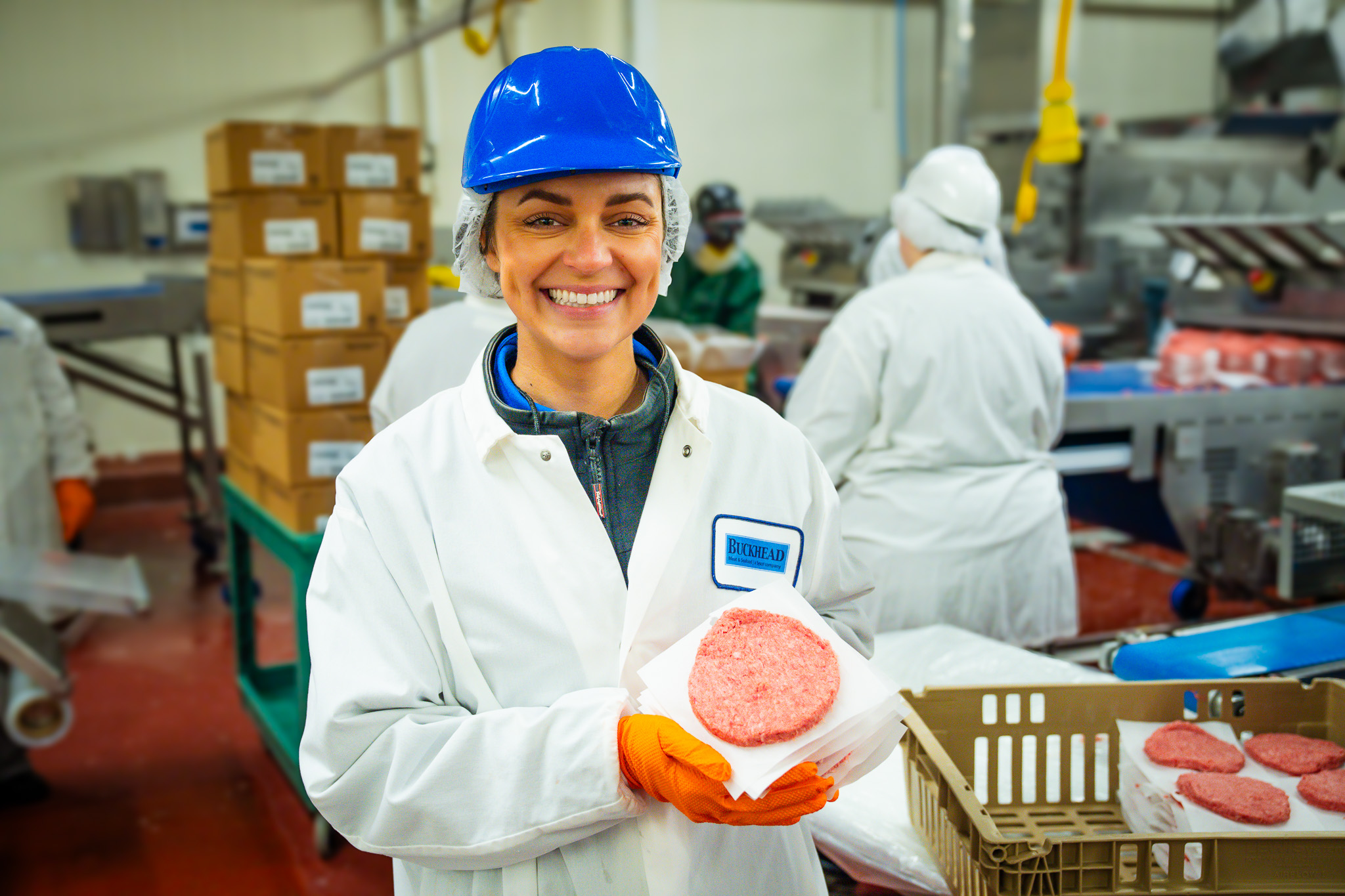 Female in hardhat holding raw burger patty in professional meat grinding facility setting.